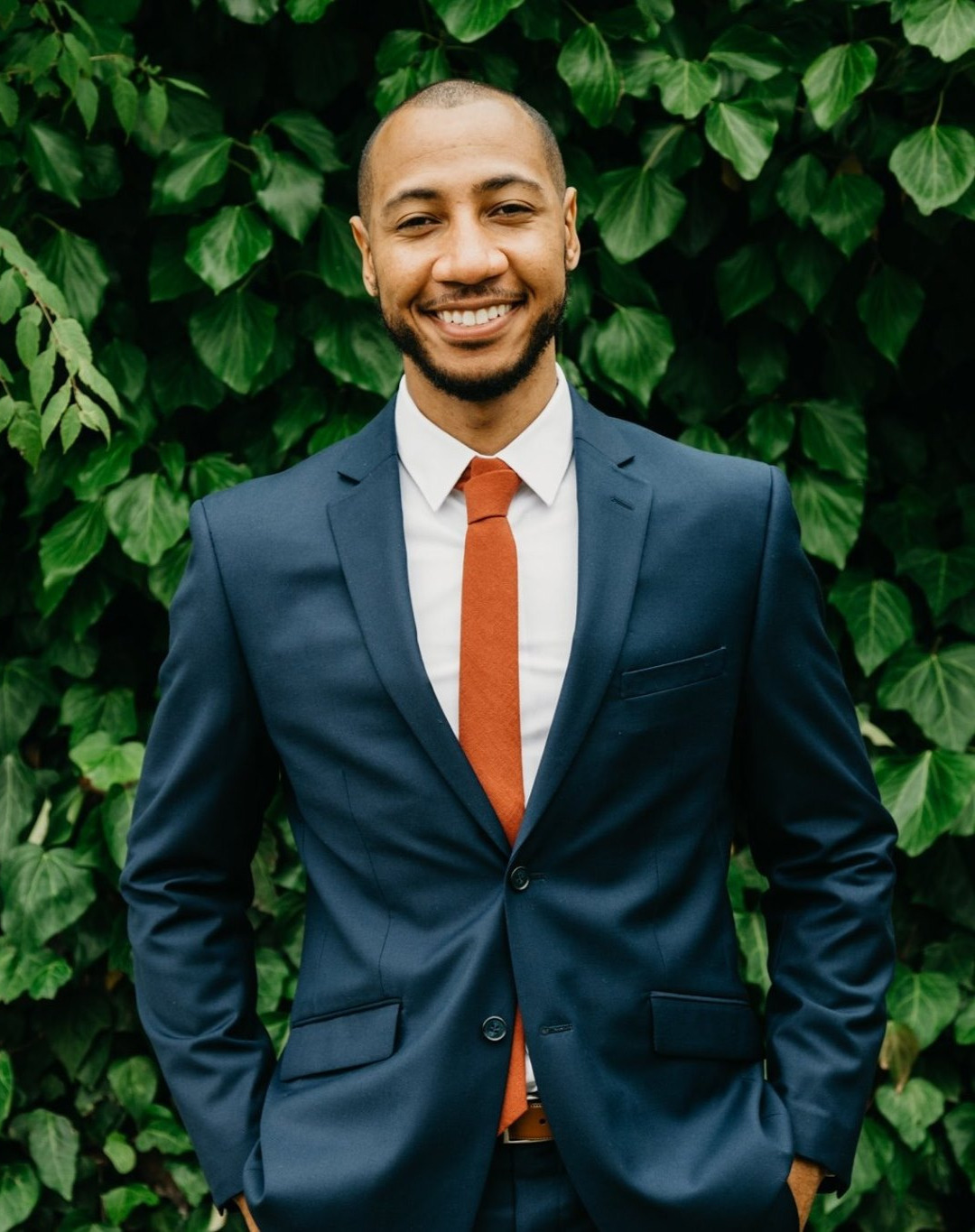 Jordan, founder of Pawtcha, smiling in a navy suit in front of a lush green ivy wall